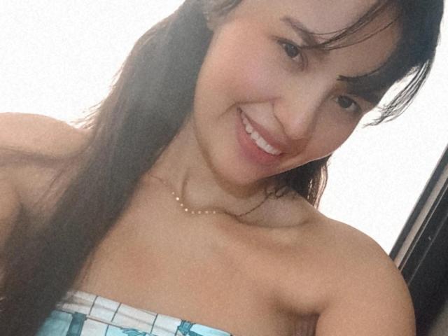 SusanKartier has long brown hair, a cute smile, and is wearing a strapless blue and white checkered top, showing her bare shoulders and a pearl necklace.