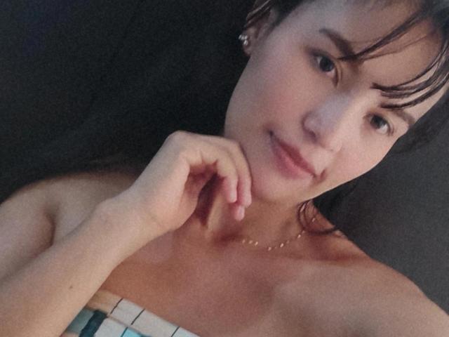SusanKartier with short brown hair, light skin, and small earrings, smiling slightly, touching her chin with her right hand, wearing a strapless top, showing her bare shoulders and chest.