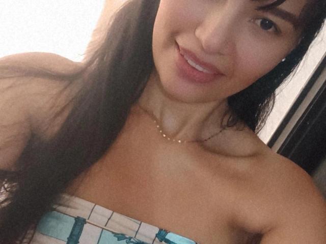 SusanKartier with long dark hair, smiling, showing off her bare shoulders and small tits in a strapless top with blue and white patterns. She's wearing a gold necklace.