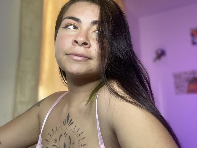 EliMartinez with long brown hair, light skin, tattoos on chest, pink bra, looking sideways with subtle smile, pinkish lighting, soft shadows, slightly messy hair, relaxed expression.