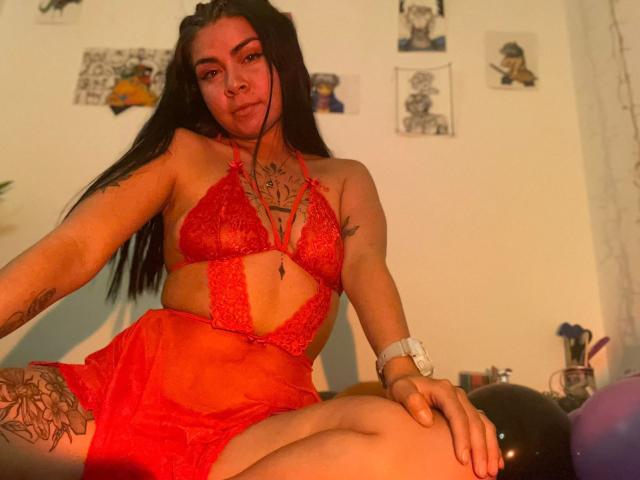 EliMartinez with long black hair, wearing red lace lingerie, showcasing her tits and tattoos, sitting on a bed with a watch on her wrist. Warm indoor lighting.