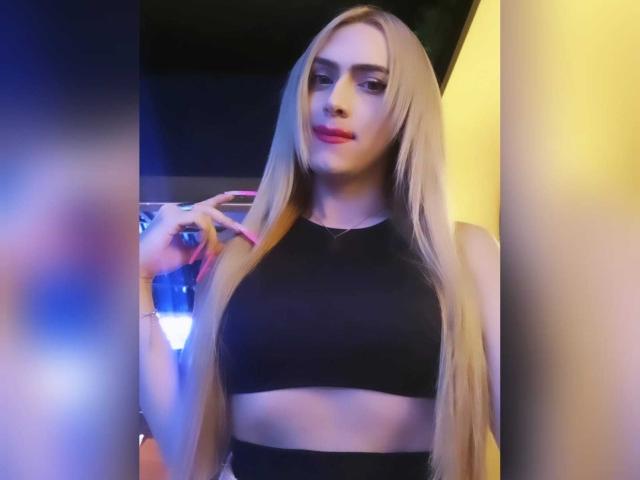 LorenaFoxy with long blonde hair, wearing a black crop top, showing her tits, pink nails, and a slight smile, holding a drink in a dimly lit room.