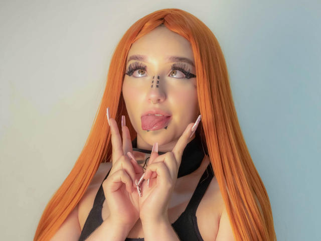 CristySanz with long orange hair, black makeup dots on face, tongue out, black choker, and long pink nails, making a "rock on" gesture. Boobs, ass not visible.