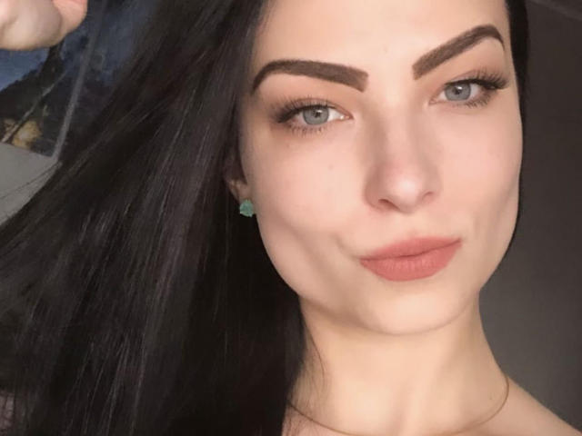 LollaHott has pale skin, dark eyebrows, blue eyes, and full pink lips with a slight smirk. She's pulling black hair to the side, wearing green earrings.