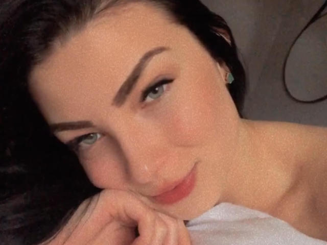 LollaHott with dark hair, green eyes, and a slight smile, resting her chin on her hand, showing her cute nose and subtle makeup. She's wearing a white top.