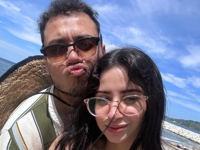 LucyAndJake with brown curly hair and glasses, pouting, stands behind Jake with black curly hair and glasses, both wearing striped shirts, on a sunny beach.