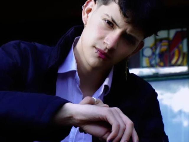 GabyThoms has a slight frown, black hair, and a nose ring, wearing a black jacket over a white shirt, with hands crossed in front of her.