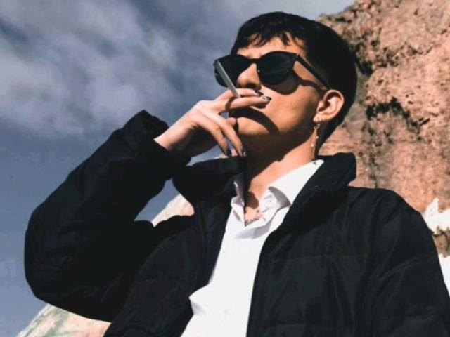 GabyThoms with dark sunglasses, black jacket, white shirt, and short black hair, holding a phone to his mouth, standing in front of a rocky background.