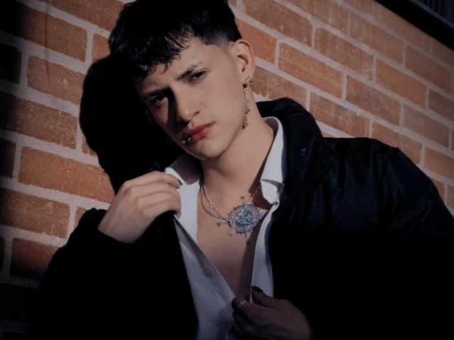 GabyThoms, with short black hair, wearing a black jacket, white shirt, and necklace, has a blue tattoo on chest, ear ring, and serious expression, against a brick wall.