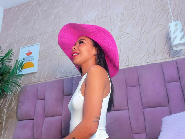 HellenSky with pink hat, white tank top, and visible arm tattoo, smiling with pink lips, sitting on purple bed, black hair, medium tits, cute, indoors.