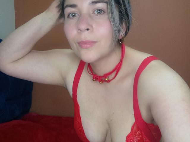 DianaAme with pale skin, blue eyes, and short gray hair, wearing a red lace bra and matching choker with gold bells, sitting on a red bed. Boobs visible.