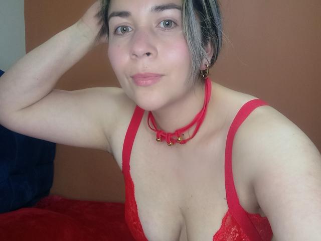 DianaAme with pale skin, blue eyes, and short gray hair, wearing red lace bra and choker with gold balls, resting arm on head, smiling slightly, showing her tits.