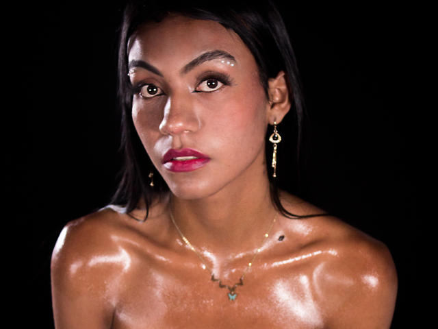 KattaleyaHorny with shiny, oiled-up tits and a serious face, wearing gold earrings and a tiny necklace, against a black background. She has dark hair and pink lipstick.