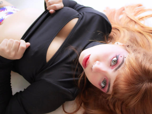 Acarolleao-hot with red hair, green eyes, and nose ring, lying down, wearing a black top that exposes her tits, holding it with both hands, looking at the camera.