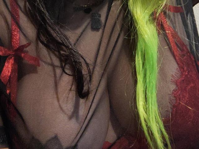 GineBralkino's sheer black mesh top with red lace and bows, showing her tits, with green and black hair in her face, and a black paw tattoo on her chest.