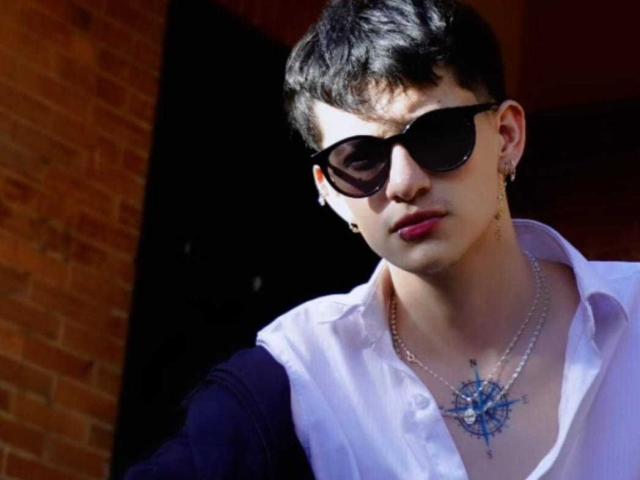 GabyThoms with short black hair, black sunglasses, white shirt, blue star necklace, earrings, and pink lipstick, looking serious with a dark background.