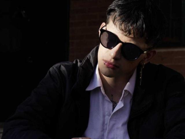 GabyThoms with short black hair, wearing dark sunglasses, black jacket, white shirt, and gold earrings, sitting in shadow, looking down, with light highlighting his face.