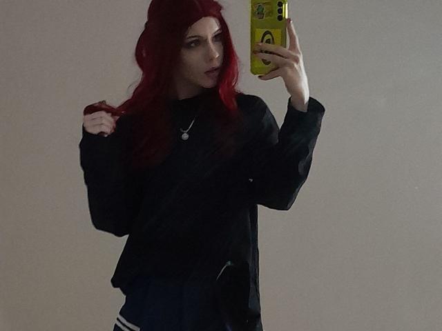 MonyBabe with red hair, black top, holding yellow phone, looking in mirror, slightly biting hair, wearing necklace, dark pants, hand on hip, casual pose, medium shot.