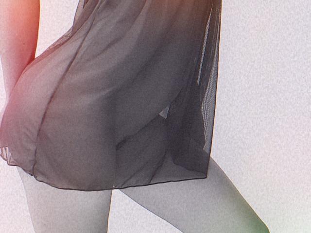 NaughtySecrets draagt een doorschijnende zwarte jurk, waardoor haar grijze onderbroek en schaamdelen zichtbaar zijn. Haar hand zit in haar jurk en ze tilt deze iets op. De afbeelding is gefocust op haar onderlichaam..