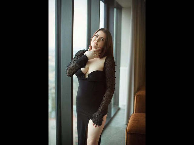 EvaLiebling-hot stands in a black dress with lace sleeves, showing her tits in a low-cut, holding her chin, next to a window, with a brown chair nearby.