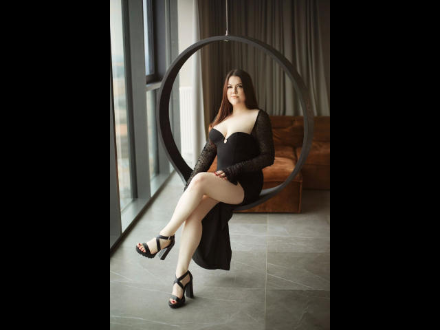 EvaLiebling-hot in a black dress with lace sleeves, sitting in a hanging chair, showing her ass, with black high heels, and fair skin, in a modern room.