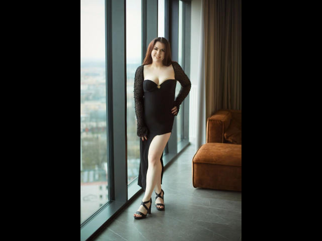EvaLiebling-hot stands in a black lace dress with a low-cut, showing her tits, and a high slit, wearing black strappy heels, in a modern room with a brown couch.