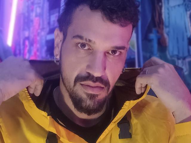 BananaX, with a curly beard and ear piercings, is pulling up a yellow jacket, looking directly at the camera with a slightly serious expression. His nose is slightly red.
