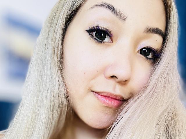 RenyLime has light blonde hair, black eyeliner, and pink lipstick. Her face is slightly tilted with a subtle smile, and she has small, barely visible tits.