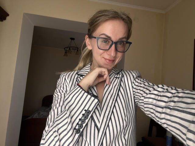 VasilisaSexy with blonde hair, blue glasses, striped shirt, and buttoned sleeves, smiling with her chin raised, showing a cute, sexy face. Her tits are slightly visible under the shirt.
