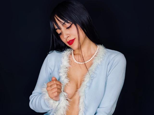 AnalisseJade with long black hair, red lipstick, and a pearl necklace, wearing a blue, furry-trimmed, open jacket, exposing her tits, against a dark background.