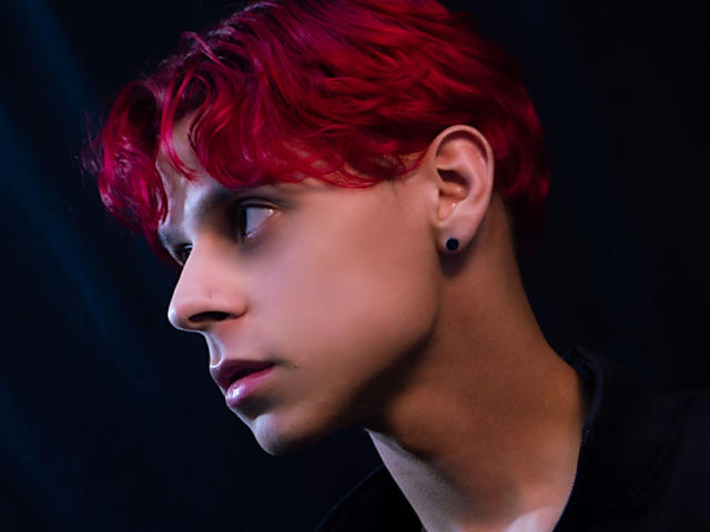 LoganEliot, avec des cheveux roux et ondulés, des bouchons d'oreille noirs et un visage sérieux, regarde vers la gauche. De légères nuances bleu clair sur sa joue, portant une chemise noire..