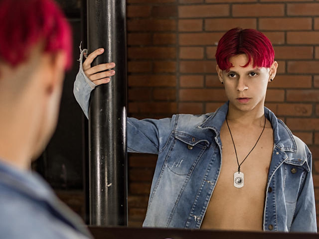LoganEliot, com cabelo ruivo, vestindo uma jaqueta jeans azul e uma placa de identificação, está em frente a um poste preto, olhando para alguém com cabelo ruivo borrado em primeiro plano..