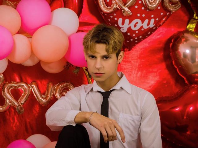 LoganEliot met blond haar, een wit shirt, een zwarte stropdas en zwarte broek, zittend, armen gekruist, met een serieuze blik, omringd door roze en witte ballonnen, glanzende rode harten en metalen bordjes met de tekst "liefde"..