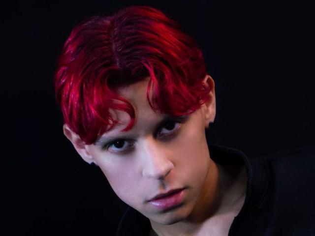 LoganEliot tiene el cabello rojo, ondulado y de estilo punk, piel pálida y una expresión seria, viste una camiseta negra, con una ligera inclinación hacia adelante, sobre un fondo oscuro..