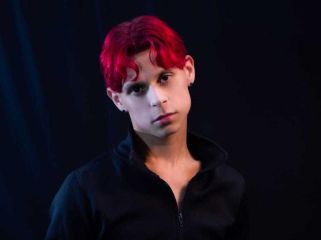 LoganEliot, con cabello rojo y rizado, lleva una sudadera negra con cremallera y tiene una expresión seria. La iluminación azul oscuro resalta su rostro, y se puede ver un pequeño arete en su oreja izquierda..
