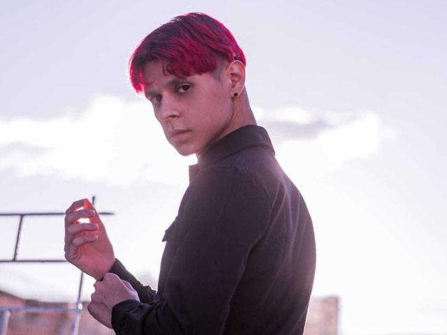 LoganEliot with red-dyed hair, black shirt, looking over shoulder, holding cigarette, serious expression, small earrings, slight shadow, soft pinkish light.