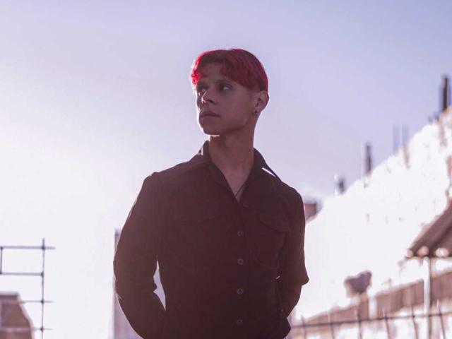 LoganEliot, con cabello rojo, vistiendo una camisa negra de botones, con las manos en los bolsillos, de pie a la luz del sol, mirando hacia un lado, en una fotografía ligeramente descolorida y con aspecto áspero..