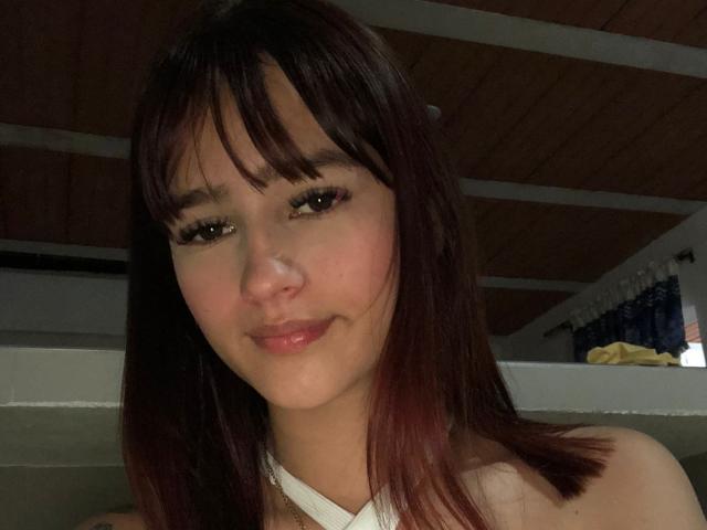 MelodyMansson with dark brown hair, bangs, and long eyelashes, smiling softly, showing her nose and pink lips, wearing a white halter top, in a dimly lit room.