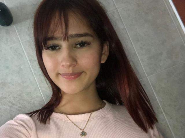MelodyMansson has red hair, brown eyes, and a cute smile. She's wearing a pink ribbed shirt and a gold necklace with a small pendant. Her tits are not visible.