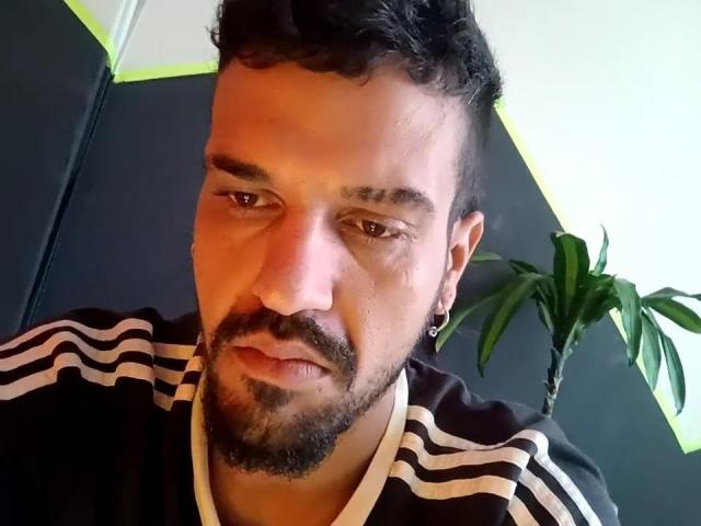 A selfie of BananaX with a serious expression, short curly black hair, small earrings, black and white striped shirt, and a stubby beard, showing a slight frown.