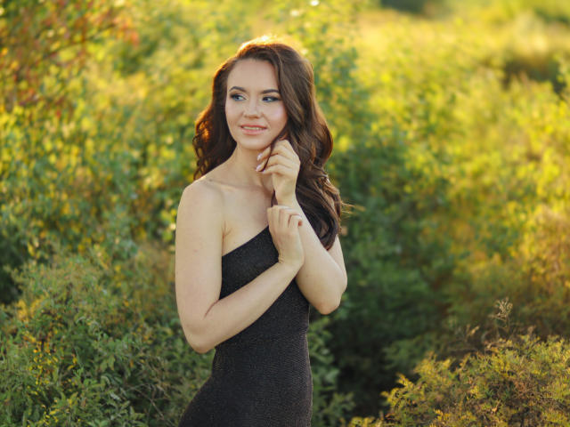 KaraCriegs with wavy brown hair, smiling, wearing a black strapless dress, standing in greenery, touching her chin with her right hand, sunlight on her face.