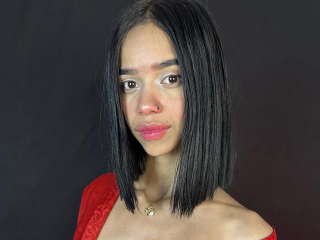 LiaBlom with black straight bob, red dress, nose ring, pink glossy lips, brown eyes, gold necklace, black background. She's looking to the side with a slight smile.