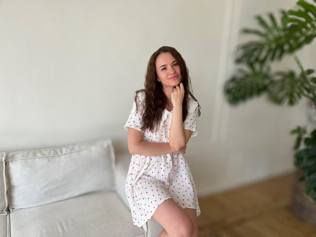 KaraCriegs with wavy brown hair, wearing a white polka-dot dress, sits on a gray couch, smiling, resting her chin on her hand, showing her ass.