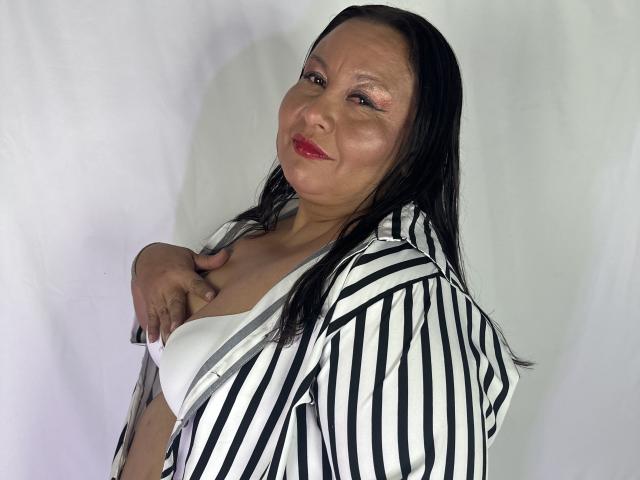 AliceShae with dark hair, red lipstick, and shiny eyeshadow, wearing a striped shirt, exposing her white bra-covered boobs, standing, looking at the camera, smiling.
