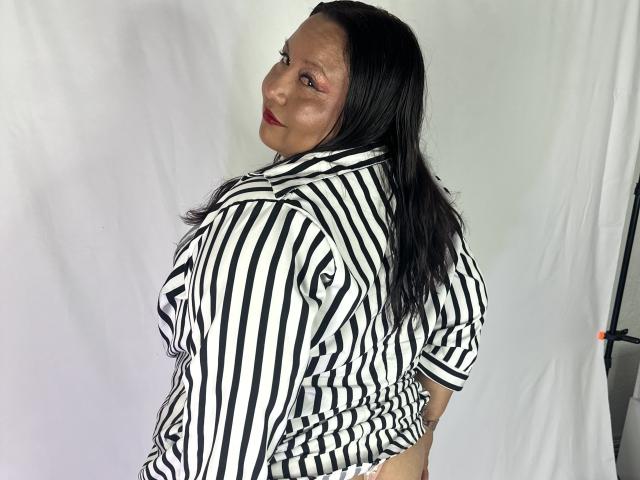 AliceShae with long black hair, wearing a black and white striped shirt, looking back over her shoulder, showing a subtle smile and red lipstick, standing in a photo.