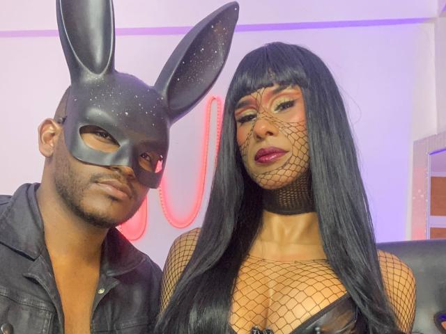 XlGoticDolly with long black hair wearing fishnet top and mask, next to bald guy in black leather jacket with bunny mask, both looking at camera. Boobs visible under net.
