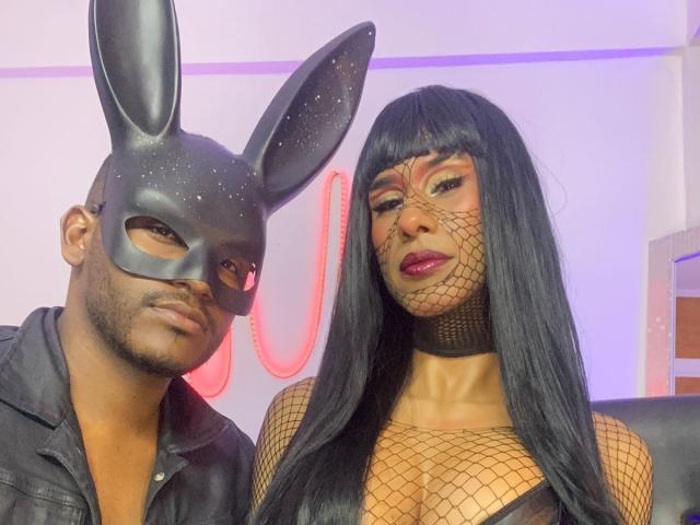 XlGoticDolly with black fishnet mask and long hair, showing her tits in fishnet, next to a bald guy in a black bunny mask and leather shirt.
