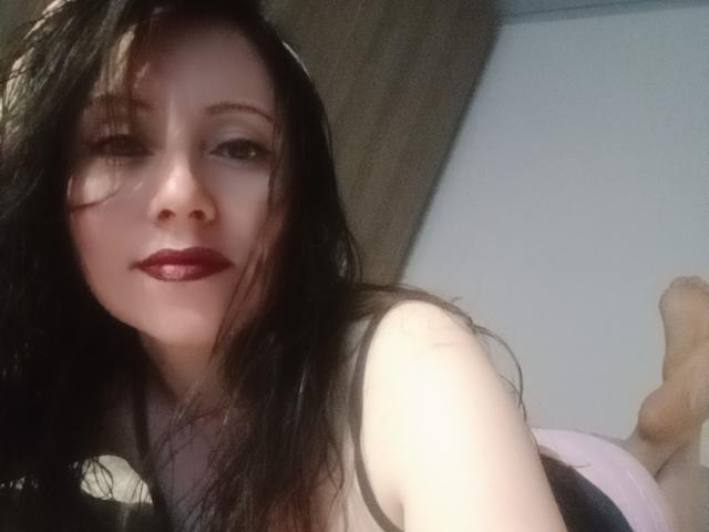 MondBlue has pale skin, dark wet hair, red lipstick, and is lying down with her tits slightly visible in a black top. Her ass is partially up in the air.