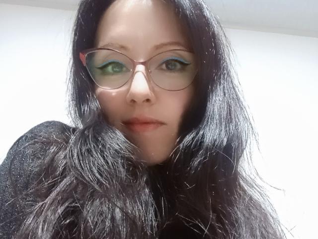 MondBlue with long black hair, green eyes, and blue eyeliner, wearing round glasses, black top, and a serious expression, looking directly at the camera.