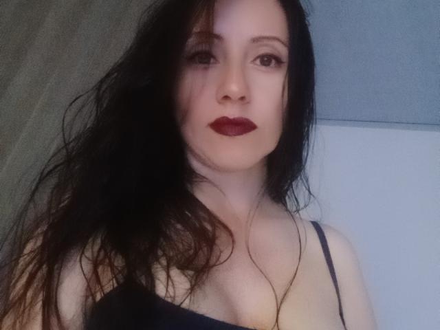 MondBlue has pale skin, dark wet hair, and red lipstick. She's wearing a black spaghetti strap top, showing her tits. She's staring into the camera with a serious look.